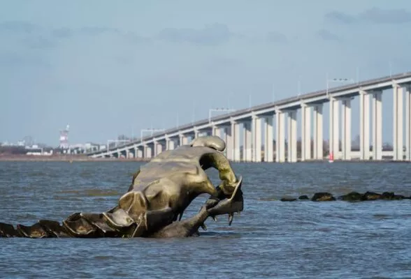Une sculpture géante en forme de squelette de serpent de mer émerge de la mer avec en arrière plan un très grand pont