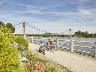 Une mère et ses deux enfants à vélo roulent sur le bord de la Loire, avec en fond le pont d'Ancenis