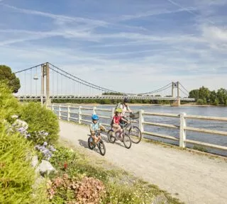 Une mère et ses deux enfants à vélo roulent sur le bord de la Loire, avec en fond le pont d'Ancenis