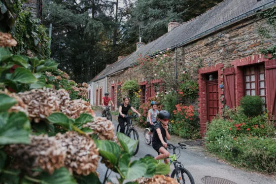 Une famille à vélo descend une côte devant une maison en pierre et des hortensias