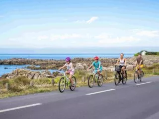 Une famille roule en vélo sur le bord de la route. Derrière eux on distingue les falaises de la côte sauvage et la mer.
