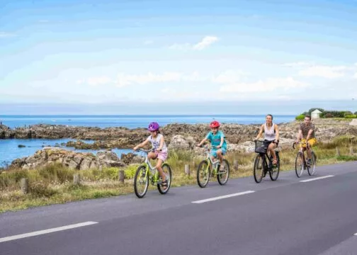 Une famille roule en vélo sur le bord de la route. Derrière eux on distingue les falaises de la côte sauvage et la mer.