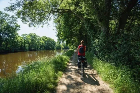 Une cycliste roule à vélo le long du canal de Nantes à Brest, à l'ombre des arbres.