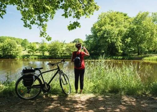 Une femme debout à côté de son vélo regarde le canal de Nantes à Brest en profitant de l'ombre des arbres