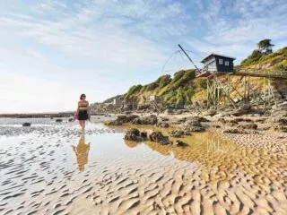 Une femme marche sur la plage à marée basse, avec derrière elle une pêcherie