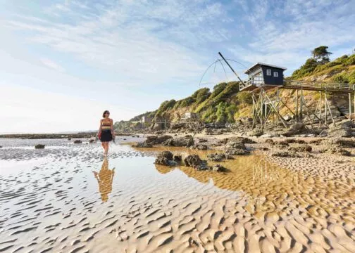 Une femme marche sur la plage à marée basse, avec derrière elle une pêcherie