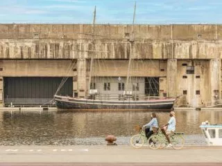 Deux cyclistes passent à vélo devant un ensemble de blockhaus en béton au bord d'une pièce d'eau. Un bateau est amarré devant.