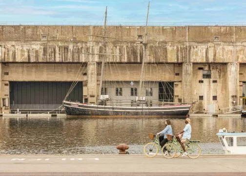 Deux cyclistes passent à vélo devant un ensemble de blockhaus en béton au bord d'une pièce d'eau. Un bateau est amarré devant.