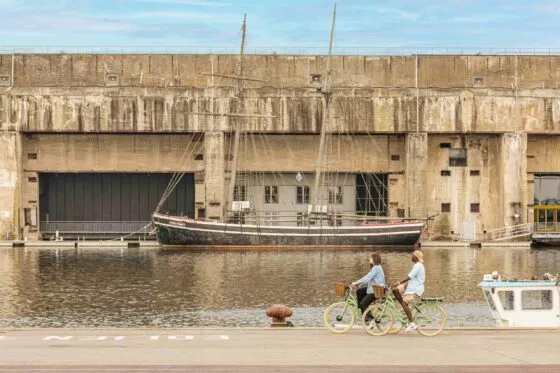 Deux cyclistes passent à vélo devant un ensemble de blockhaus en béton au bord d'une pièce d'eau. Un bateau est amarré devant.