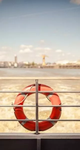 Vue d'un bateau s'éloignant de Nantes, avec au premier plan le bastingage et les bouées de sauvetage qui y sont accrochées. Au second plan, on distingue l'île de Nantes, reconnaissable à la Grue Titan jaune.