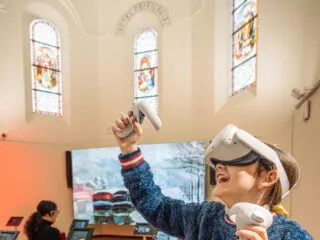 Une adolescente portant un casque de réalité virtuelle et tenant des manettes dans ses deux mains expérimente en riant. Derrière elle, une femme regarde une tablette. Elles sont dans une ancienne chapelle dont on peut voir les vitraux, au dessus de leurs têtes.