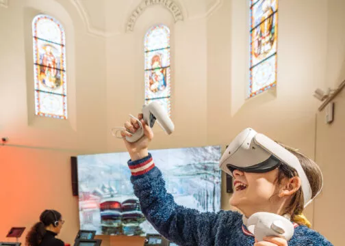 Une adolescente portant un casque de réalité virtuelle et tenant des manettes dans ses deux mains expérimente en riant. Derrière elle, une femme regarde une tablette. Elles sont dans une ancienne chapelle dont on peut voir les vitraux, au dessus de leurs têtes.