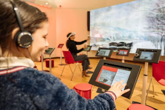 Une jeune fille consulte une tablette numérique posée sur pied. Elle a un casque audio sur la tête et navigue sur la tablette pour découvrir les œuvres présentées. Au second plan, une femme utilise un casque de réalité virtuelle. Un grand écran affiche une œuvre picturale dans le fond.