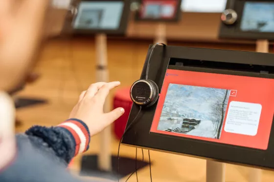 Gros plan sur une tablette numérique installée sur pied et affichant une œuvre picturale sur son écran. La main d'une jeune fille se tend vers le casque accroché sur le côté.