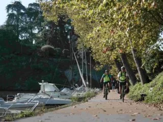 Deux cyclistes pédalent sur une piste cyclable au bord de l'Erdre. A gauche, des bateaux amarrés au bord de l'eau, à droite des arbres qui commencent à perdre leurs feuiles