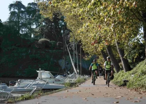 Deux cyclistes pédalent sur une piste cyclable au bord de l'Erdre. A gauche, des bateaux amarrés au bord de l'eau, à droite des arbres qui commencent à perdre leurs feuiles