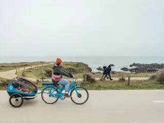 Un cycliste tirant une remorque dans laquelle est assit un enfant pédale sur une piste cyclable qui longe la mer et les rochers.