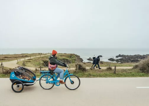 Un cycliste tirant une remorque dans laquelle est assit un enfant pédale sur une piste cyclable qui longe la mer et les rochers.