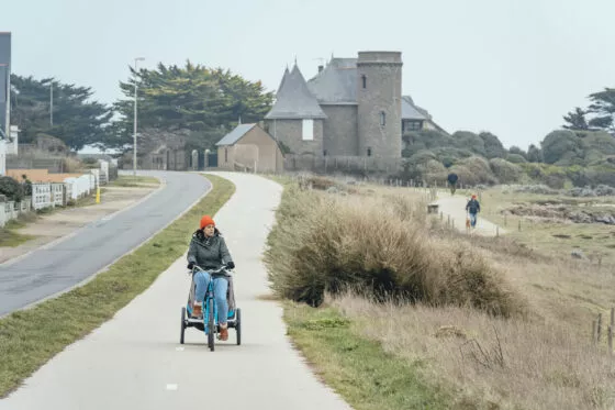 Un cycliste tirant une remorque où est installé un enfant pédale sur une piste cyclable qui borde le chemin des douaniers et longe la côte sauvage. Derrière lui, des maisons en pierres se détachent d'un ciel gris.