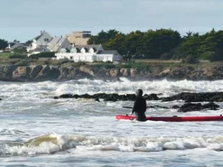 Un surfeur portant une combinaison intégrale entre dans l'eau agitée avec son surf. Il est entouré de rochers noirs qui sortent de l'eau. Derrière lui on peut voir le sentier des douaniers qui longe la mer, des maisons et un ancien Blokhaus.