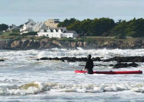 Un surfeur portant une combinaison intégrale entre dans l'eau agitée avec son surf. Il est entouré de rochers noirs qui sortent de l'eau. Derrière lui on peut voir le sentier des douaniers qui longe la mer, des maisons et un ancien Blokhaus.