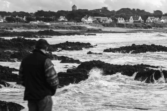 Un homme de dos, dans une doudoune, regarde la mer s'écraser sur les rochers de la côte sauvage en contrebas. au loin, la côté continue, avec des maisons et des arbres.