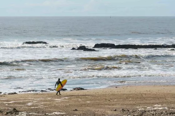 Un surfeur en combinaison intégrale marche sur la plage en direction de la mer et des vagues avec son surf sous le bras.