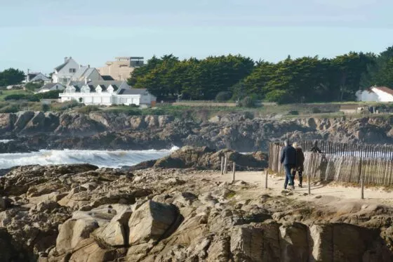 Couple marchant sur le sentier des douaniers, qui borde la côte sauvage et ses rochers. La mer se distingue dans les creux et on voit au loin des maisons et un blockhaus