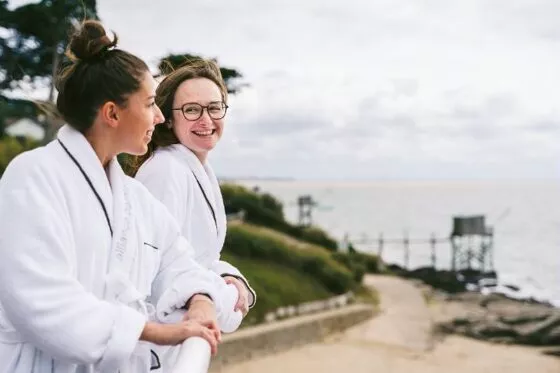 Deux jeunes femmes souriantes discutent face à l'océan depuis le balcon de la thalasso de Pornic.