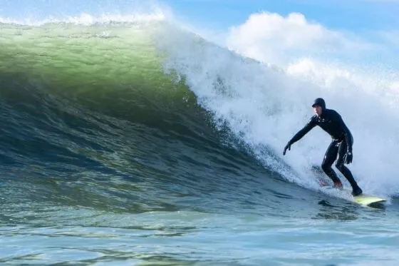 Un homme en combinaison de surf intégrale surfe sur une vague, debout sur sa planche. Derrière lui, la vague se brise en un gerbe d'écume