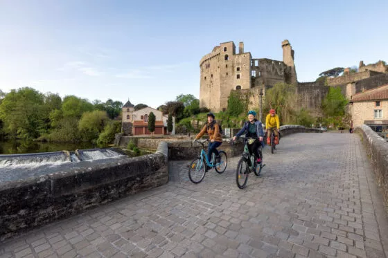 Deux cyclistes roulent sur un pont en pierre enjambant une rivière bordée d'arbres. Derrière eux s'élèvent les ruines d'un château médiéval.