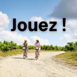 Deux femmes en tenues d'été pédalent sur des vélos. Elles roulent sur une route en terre, entre des rangs de vignes.