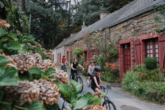 des cyclistes passant devant une maison en pierres à volets rouges