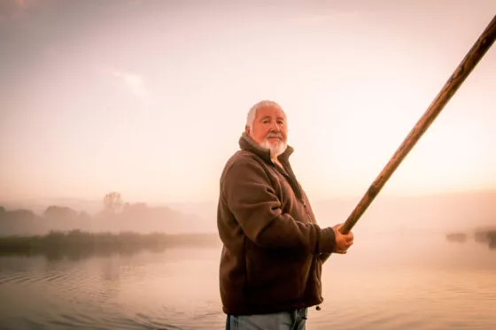 Un homme tient une perche à la main, dans un paysage d'eau au soleil couchant
