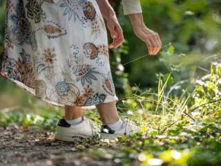 La main d'une femme habillée d'une robe d'été fleurie et chaussée de baskets se tend vers des fleurs dans un sous bois